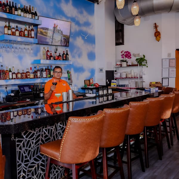 A bartender in an orange shirt holds a glass of red wine behind a bar with brown leather stools.